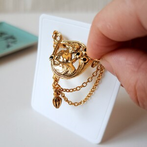 May include: A gold-toned brooch featuring a central relief figure of a person surrounded by floral motifs with small black accents. The piece includes two delicate hanging gold chains and a small dangling charm attached to the bottom edge.