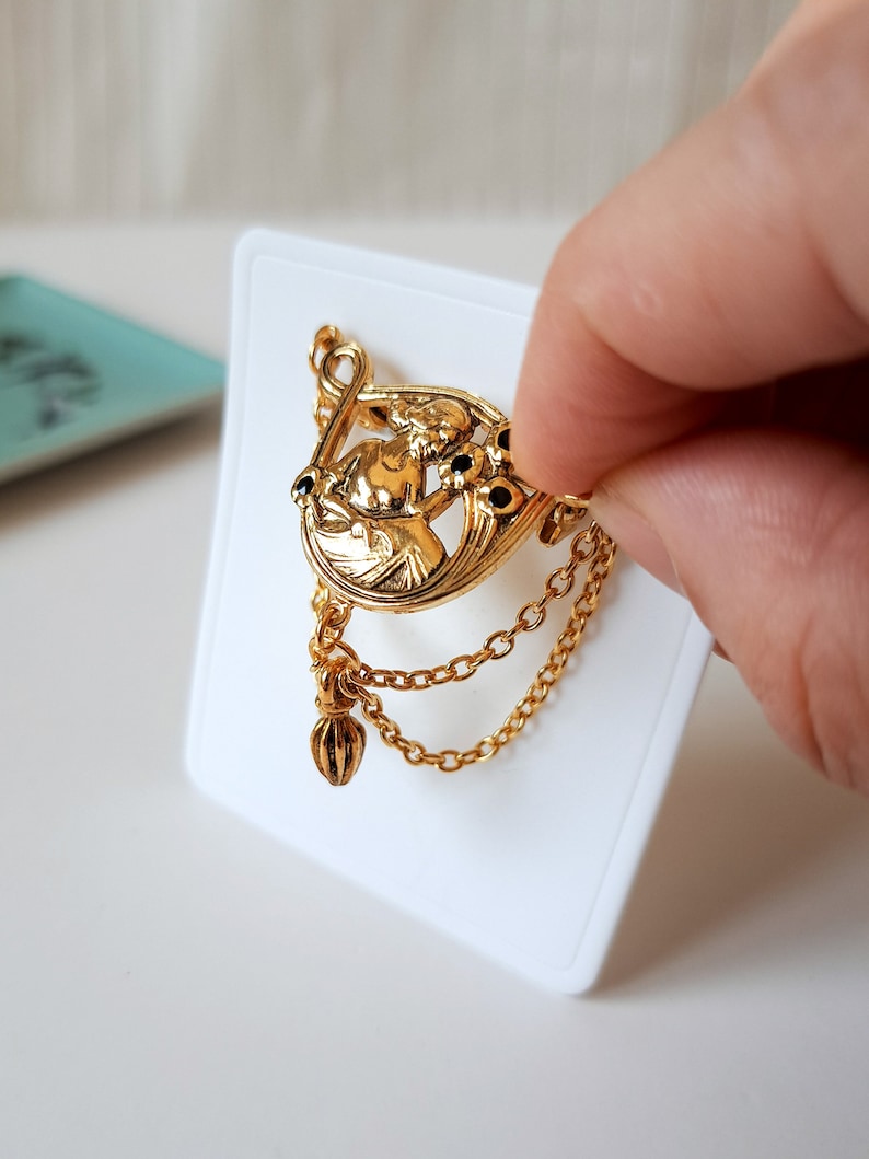 May include: A gold-toned brooch featuring a central relief figure of a person surrounded by floral motifs with small black accents. The piece includes two delicate hanging gold chains and a small dangling charm attached to the bottom edge.