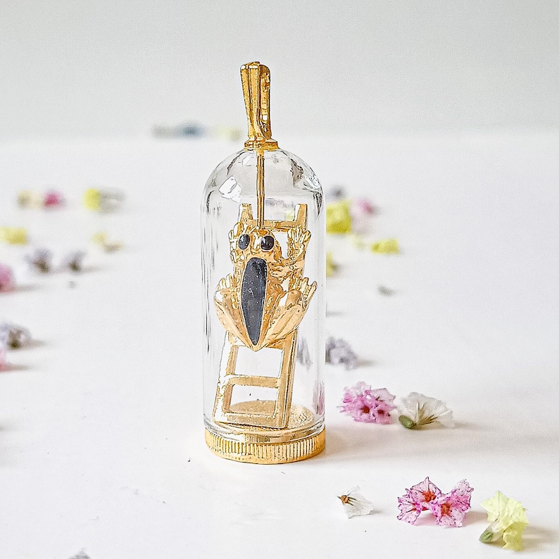May include: A gold pendant featuring a small frog figurine climbing a ladder inside a glass dome. The frog has black eyes and a black body.