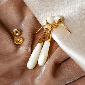 Può includere: Un paio di orecchini dorati con accenti di perle bianche a forma di lacrima. Gli orecchini sono su uno sfondo di tessuto marrone e bianco.