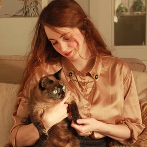 May include: A long-sleeved, champagne-colored satin button-down shirt featuring a gold-toned collar chain accessory. The accessory consists of two circular black and gold pins attached to the collar points, connected by a draped gold metal chain.