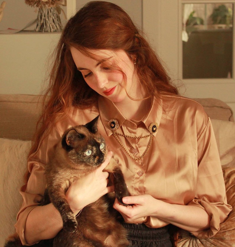 May include: A long-sleeved, champagne-colored satin button-down shirt featuring a gold-toned collar chain accessory. The accessory consists of two circular black and gold pins attached to the collar points, connected by a draped gold metal chain.