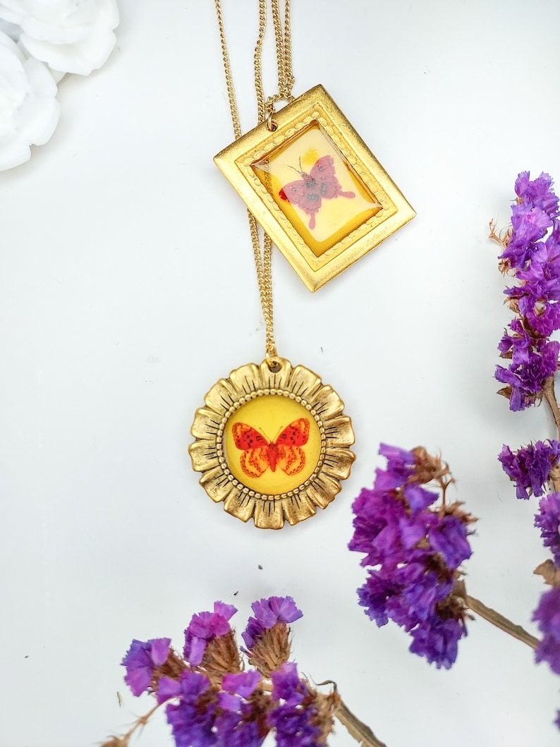 May include: Gold chain necklace with two pendants featuring butterflies encased in glass. The pendants are framed in gold with a detailed design. The butterflies are red and orange.