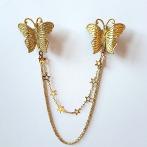 May include: A gold-toned metal cardigan clip featuring two butterfly-shaped clasps connected by two distinct chains. One chain is a simple twisted rope style, while the other is a delicate link chain adorned with small, flat, five-pointed star charms.