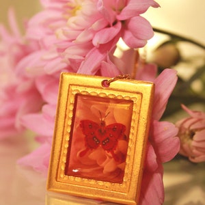 May include: A gold-framed pendant with a red butterfly and pink flowers inside a clear resin. The pendant is surrounded by pink flowers.