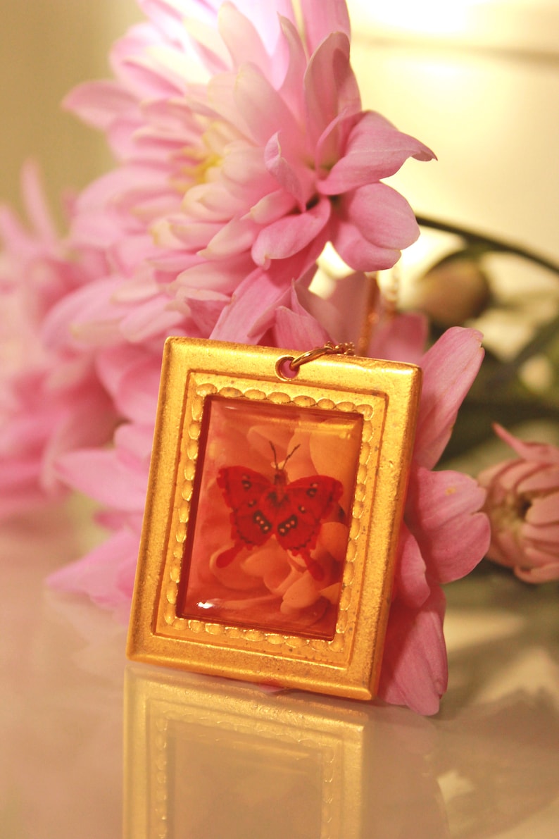 May include: A gold-framed pendant with a red butterfly and pink flowers inside a clear resin. The pendant is surrounded by pink flowers.