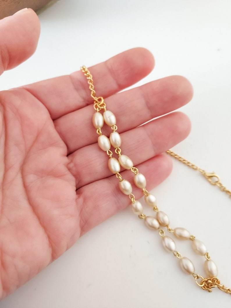 May include: A gold chain necklace with white oval-shaped beads. The necklace is on a hand.