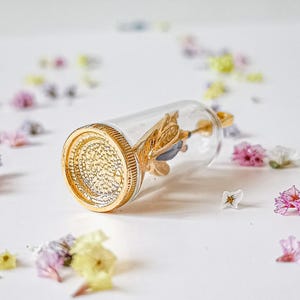 May include: A gold-toned glass bottle pendant with a gold-colored butterfly inside. The bottle is clear and has a textured surface. The pendant is lying on a white surface with scattered dried flowers.
