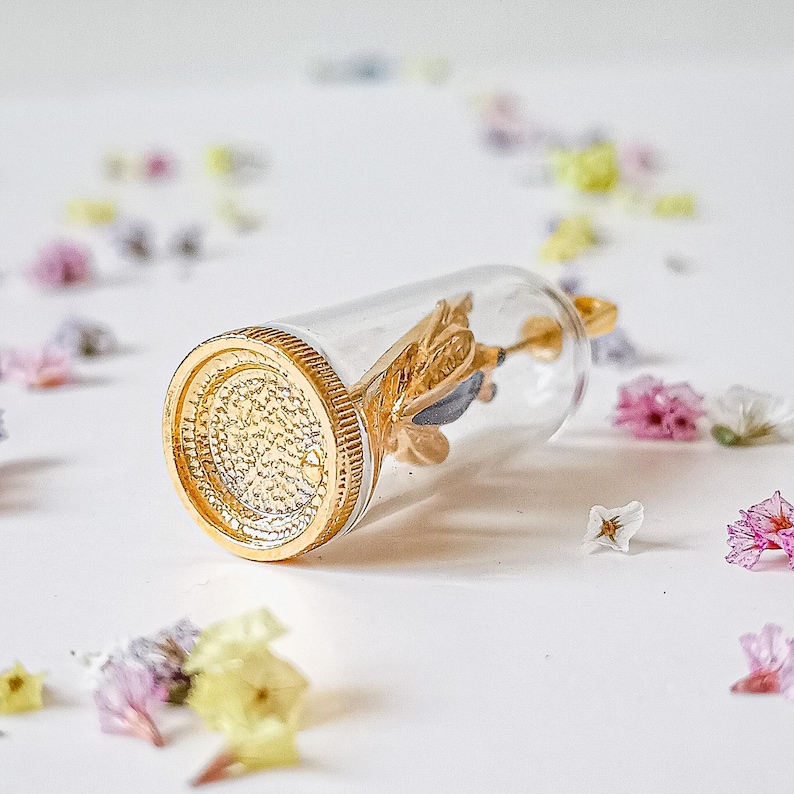 May include: A gold-toned glass bottle pendant with a gold-colored butterfly inside. The bottle is clear and has a textured surface. The pendant is lying on a white surface with scattered dried flowers.