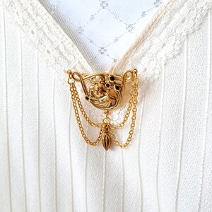 May include: A gold-toned metal brooch featuring a relief of a figure surrounded by small black stones, accented with two draped gold chains and a central dangling charm, pinned to a white fabric garment with lace trim and vertical textured stripes.