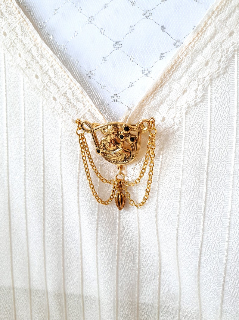 May include: A gold-toned metal brooch featuring a relief of a figure surrounded by small black stones, accented with two draped gold chains and a central dangling charm, pinned to a white fabric garment with lace trim and vertical textured stripes.