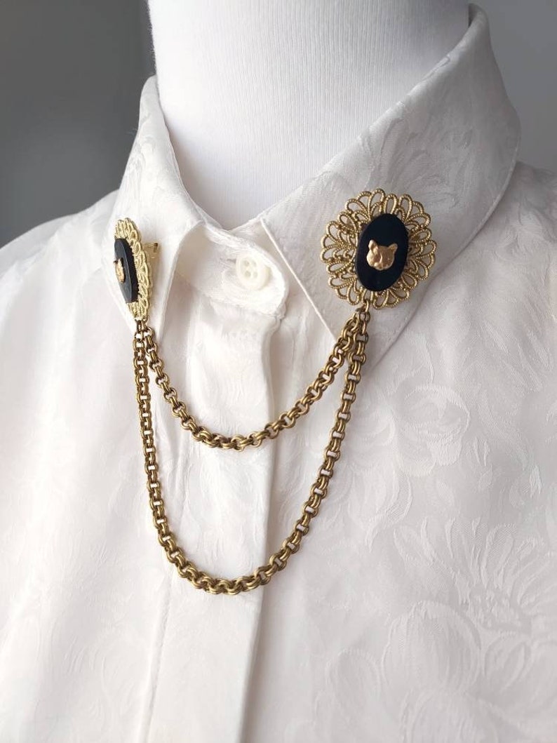 May include: A gold-toned metal collar pin set featuring two oval black cabochons with a small gold cat head emblem on each. The two pins are connected by two draped gold-toned metal chains, attached to a white button-down shirt with a floral pattern.