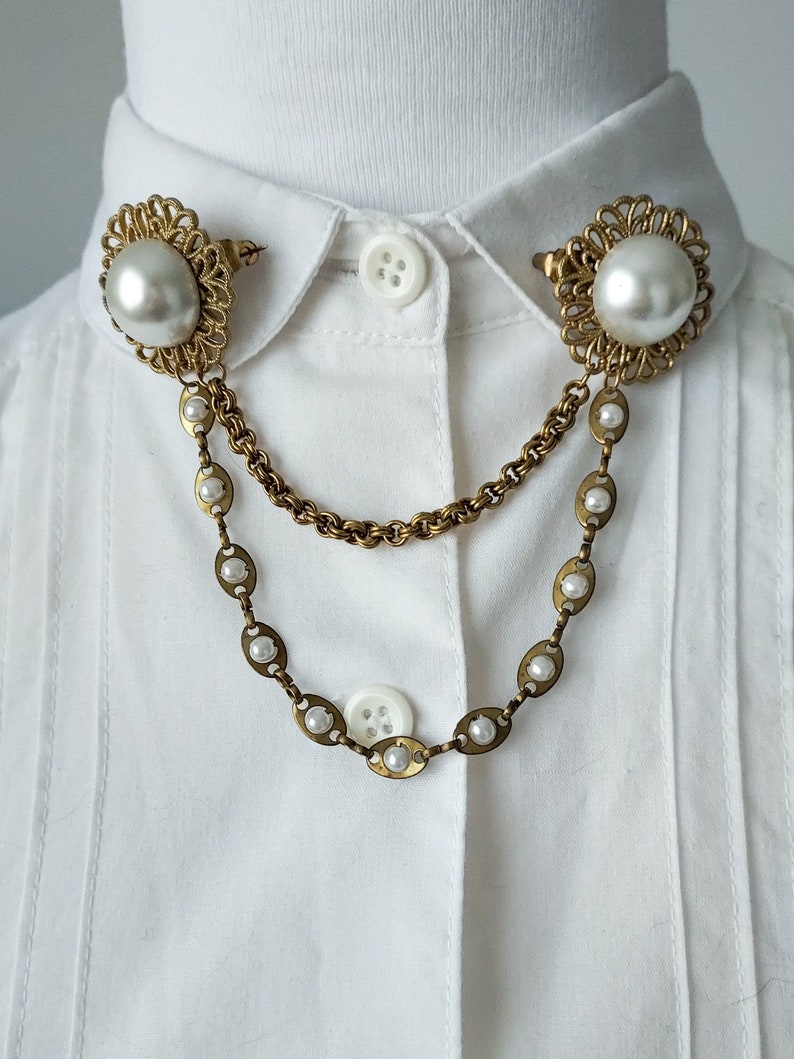 May include: A gold-toned collar pin set featuring two round faux pearl studs with intricate filigree borders. The studs are connected by two draped chains, one simple link chain and one longer chain accented with small, evenly spaced faux pearls.