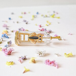 May include: A gold-colored metal frog figurine inside a clear glass bottle with a gold-colored metal stopper. The bottle is lying on a white surface with scattered flower petals.