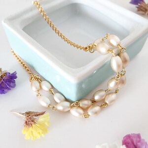 May include: A delicate gold chain bracelet with three strands of oval-shaped pearls. The bracelet is displayed on a light blue ceramic dish.