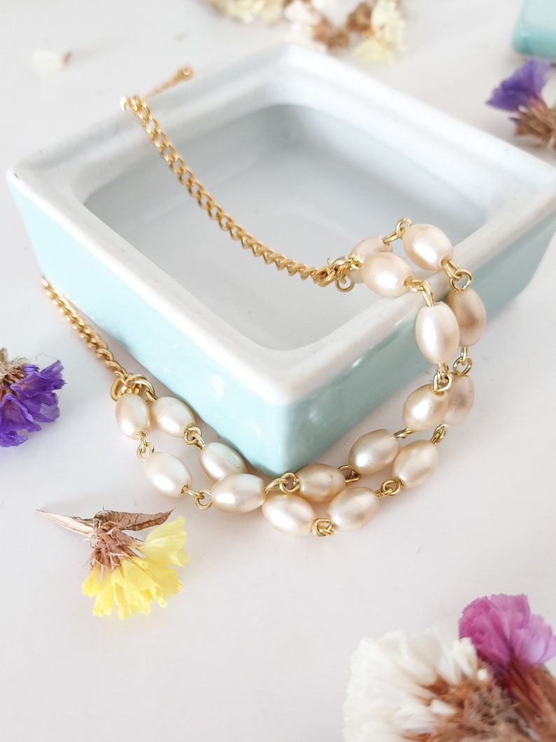 May include: A delicate gold chain bracelet with three strands of oval-shaped pearls. The bracelet is displayed on a light blue ceramic dish.