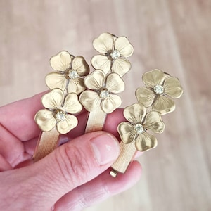 Może przedstawiać: Trzy złote spinki do włosów z kwiatowym wzorem. Każda spinka ma dwa kwiaty z kryształem w środku.