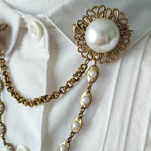 May include: A gold-toned brooch featuring a large central faux pearl surrounded by an intricate filigree frame. Two dangling chains are attached to the brooch, one a textured link chain and the other a series of oval links inlaid with small faux pearls.