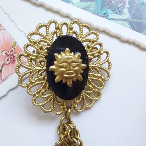 May include: A gold-tone metal brooch featuring a filigree circular frame surrounding a central oval black cabochon. A gold-tone sun face with rays is mounted on the black oval. Two gold-tone metal chains hang from the bottom of the brooch.