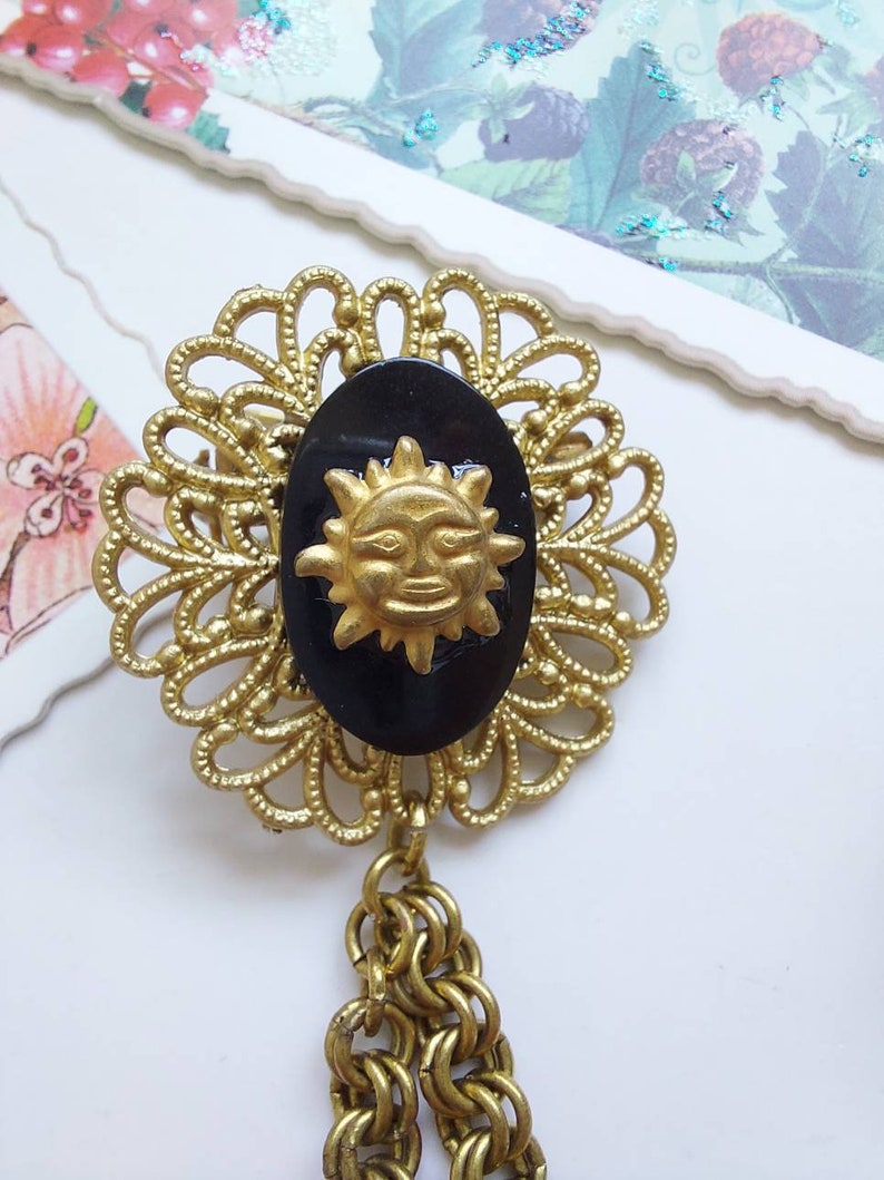 May include: A gold-tone metal brooch featuring a filigree circular frame surrounding a central oval black cabochon. A gold-tone sun face with rays is mounted on the black oval. Two gold-tone metal chains hang from the bottom of the brooch.