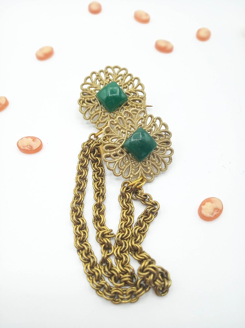 May include: A gold-tone metal brooch featuring two circular filigree discs, each centered with a square green stone. Three draped gold-tone chains hang from the lower disc. Several small, oval, peach-colored cameo cabochons are scattered nearby.