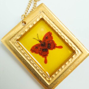 May include: A gold pendant necklace with a red butterfly encased in a yellow resin frame. The frame has a gold border with a decorative pattern.