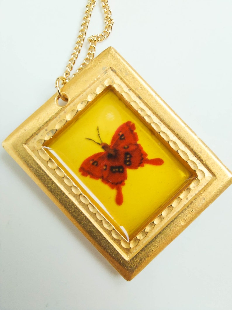 May include: A gold pendant necklace with a red butterfly encased in a yellow resin frame. The frame has a gold border with a decorative pattern.