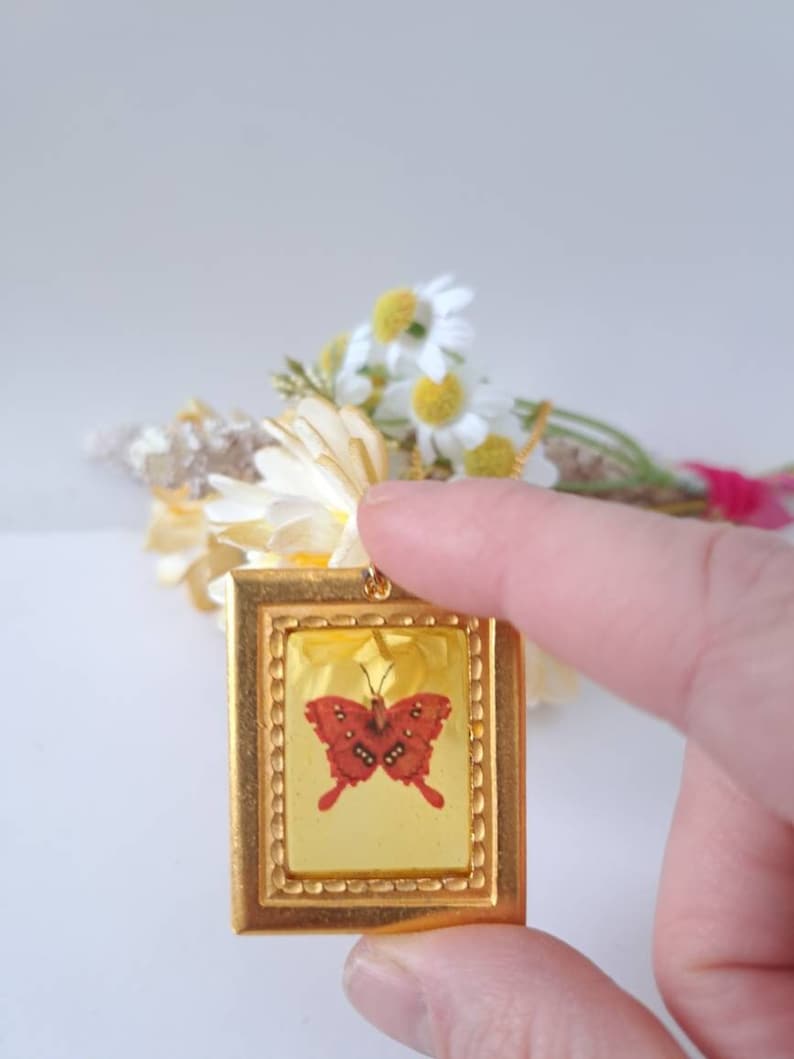 May include: A gold-toned pendant featuring a framed image of a red butterfly with black markings. The pendant is held by a finger against a white background.