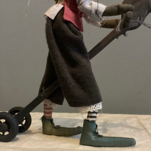 Op de afbeelding: Een handgemaakte pop met een grillig ontwerp. De pop draagt een donkerbruine broek, een rood vest en een wit overhemd. De voeten van de pop zitten in groene schoenen en ze houdt een donkergrijze kar met zwarte wielen vast. De sokken van de pop hebben rood-witte strepen.
