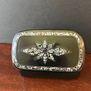 May include: A black rectangular box with a decorative star design made of iridescent shell pieces. The box has a raised border around the lid.