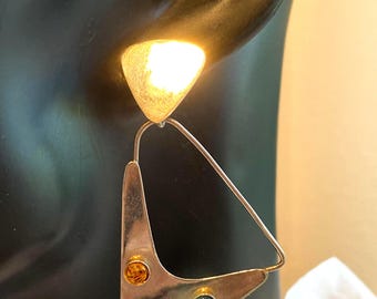 May include: A silver-toned dangle earring featuring a triangular metal frame with a textured triangular stud top. The lower frame is adorned with three small, round, bezel-set cabochon stones in amber, dark green, and deep red colors.