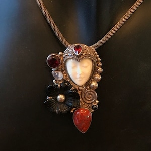 May include: A silver chain necklace with a pendant featuring a carved ivory face with red and black gemstones. The pendant is surrounded by silver filigree and a red coral stone.