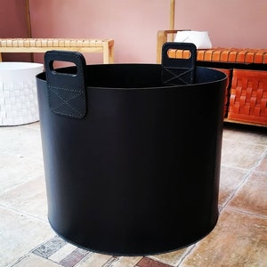May include: A cylindrical black leather storage basket with two stitched handles. The handles are attached to the exterior of the basket. The basket is sitting on a tiled floor. Other furniture is visible in the background.