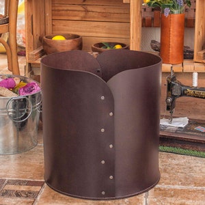 May include: A cylindrical dark brown leather storage bin featuring a vertical overlapping seam secured with six silver-toned metal rivets. The structure has a smooth, matte finish and a curved top edge, standing upright on a tiled floor surface.