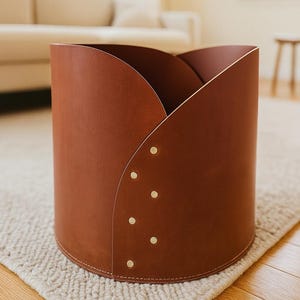 May include: A cylindrical storage bin crafted from smooth, reddish-brown leather. The top edge features a curved, overlapping petal-like design secured by a vertical row of six small, circular brass-colored metal rivets along the front seam.