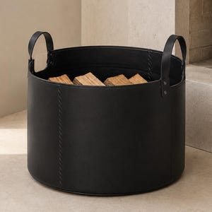 May include: A cylindrical black storage basket with two vertical handles attached by rivets. The basket features visible stitched seams along the sides and is filled with several pieces of light brown firewood logs inside the main compartment.