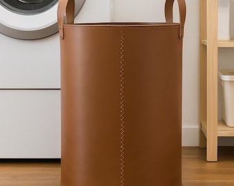 May include: A cylindrical brown laundry hamper featuring a vertical stitched seam running down the center and two matching brown handles attached to the top rim. The hamper has a smooth, solid-colored exterior and a deep, open-top design.