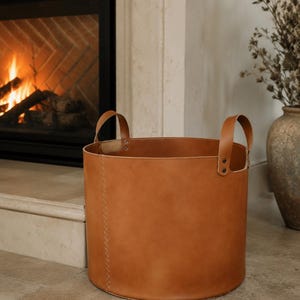May include: A cylindrical tan leather storage basket featuring two matching leather handles attached with metal rivets. The basket has a visible vertical stitched seam running down the side and a smooth, matte finish on the exterior surface.
