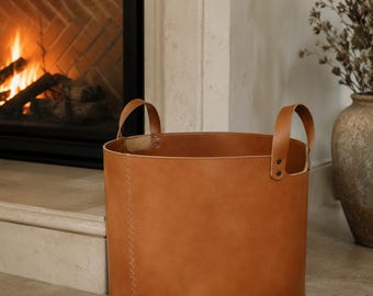 Puede incluir: Una cesta de almacenamiento cilíndrica de cuero color canela con dos asas de cuero a juego sujetas con remaches metálicos. La cesta tiene una costura vertical visible en el lateral y un acabado mate liso en la superficie exterior.