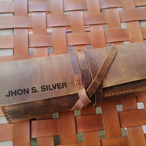 以下が含まれることがあります： ストラップ留めの茶色の革製のロールアップケース。ケースには「JHON S. SILVER」という名前が黒文字で刻印されています。