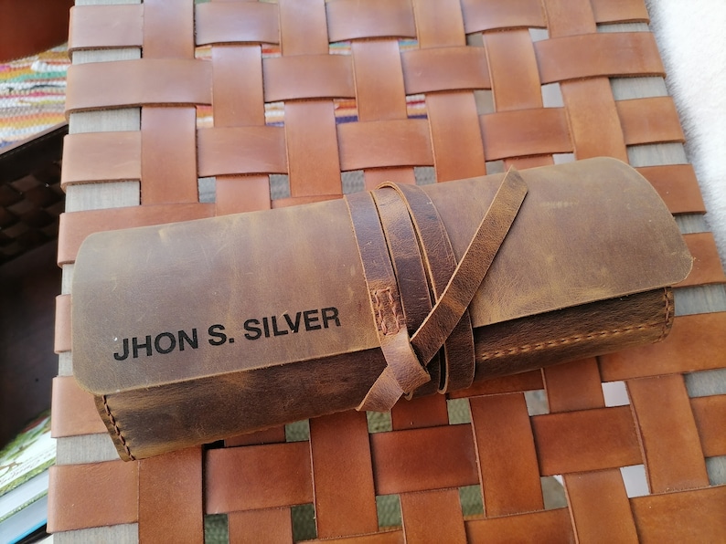 以下が含まれることがあります： ストラップ留めの茶色の革製のロールアップケース。ケースには「JHON S. SILVER」という名前が黒文字で刻印されています。