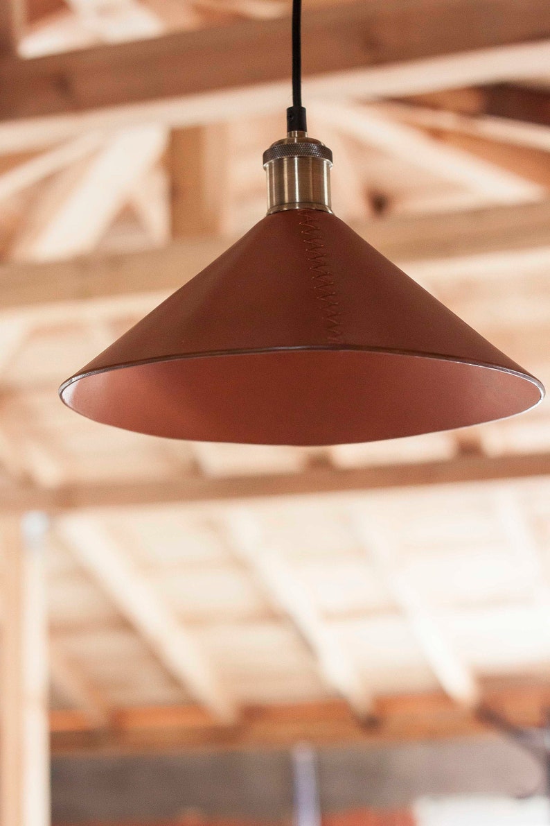 May include: A cone-shaped pendant light fixture featuring a smooth, dark brown leather shade with visible stitching along the side. The lamp is suspended by a black cord and includes a metallic, textured brass-toned socket at the top.