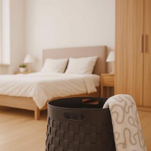 K&ouml;nnte beinhalten: Ein dunkelbrauner geflochtener Korb mit einer cremefarbenen Decke mit grauem, abstraktem Muster. Der Korb hat zwei Griffe und steht auf einem hellen Holzboden. Die Szene spielt in einem Schlafzimmer.