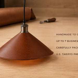 May include: A conical pendant lamp shade crafted from brown leather with visible stitching, suspended by a black cord from a metallic fixture. Text on the right reads: HANDMADE TO ORDER, UP TO 7 BUSINESS DAYS, CAREFULLY PACKED, U.S. TARIFFS PREPAID.