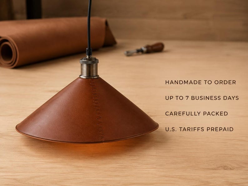 May include: A conical pendant lamp shade crafted from brown leather with visible stitching, suspended by a black cord from a metallic fixture. Text on the right reads: HANDMADE TO ORDER, UP TO 7 BUSINESS DAYS, CAREFULLY PACKED, U.S. TARIFFS PREPAID.