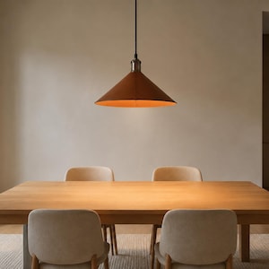 May include: A conical pendant light fixture with a matte rust-colored exterior and a warm orange interior finish, suspended by a thin black cord above a rectangular light-toned wooden dining table with four matching upholstered chairs.