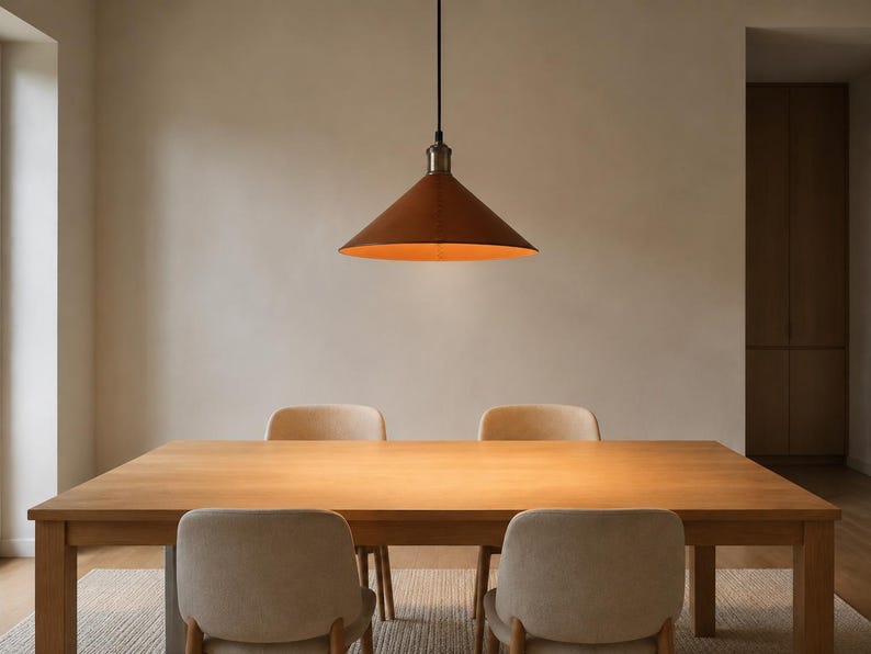 May include: A conical pendant light fixture with a matte rust-colored exterior and a warm orange interior finish, suspended by a thin black cord above a rectangular light-toned wooden dining table with four matching upholstered chairs.