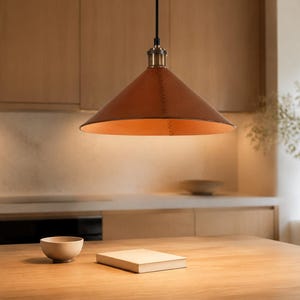 May include: A conical pendant light fixture featuring a smooth, tan leather shade with visible stitched detailing along the side. The lamp is suspended by a black cord and includes a metallic brass-toned hardware fitting at the top center.
