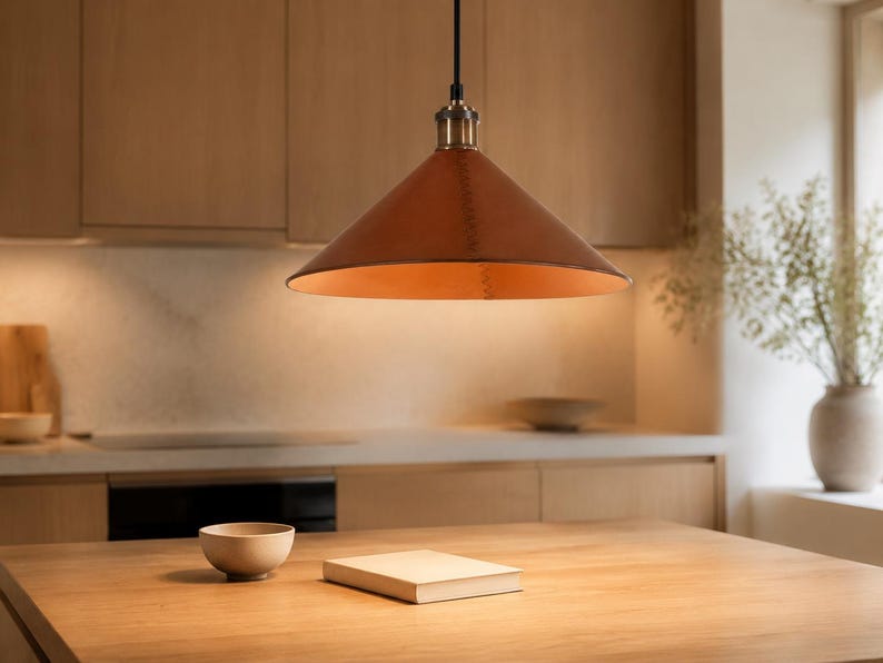 May include: A conical pendant light fixture featuring a smooth, tan leather shade with visible stitched detailing along the side. The lamp is suspended by a black cord and includes a metallic brass-toned hardware fitting at the top center.