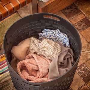 Puede incluir: Una cesta de almacenamiento redonda de tejido gris oscuro con asas integradas, llena de una variedad de textiles plegados que incluyen una tela rosa texturizada, una tela beige estampada, una tela azul con corazones y una pieza de tela gris lisa.